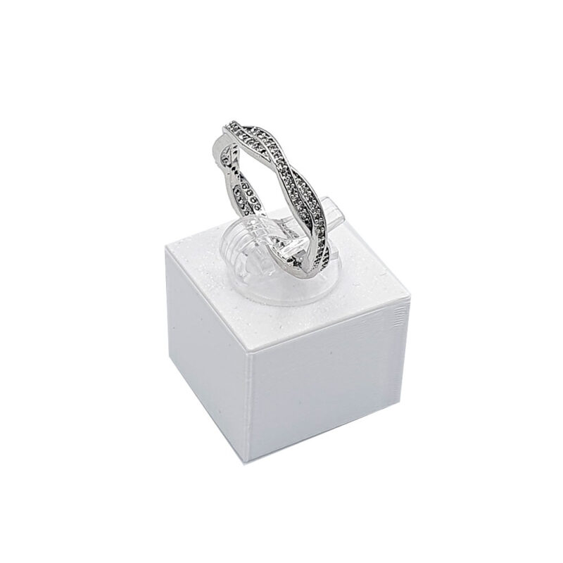 Βεράκι πλεκτό 2 - bijouxbijoux Silver ring with small dark stones on a clear display stand atop a white jewelry box, product photo, studio lighting.