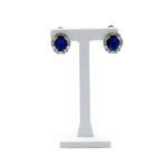 Blue oval sapphire stud earrings with a halo of small diamonds on a silver stand.