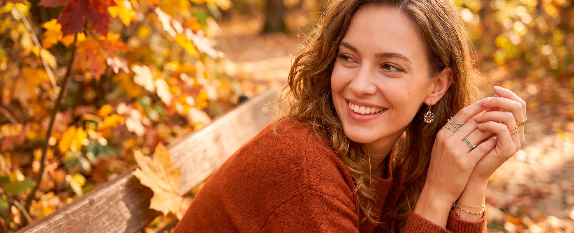 Smiling woman with wavy hair in an orange sweater sits on a wooden bench in a park surrounded by autumn leaves.